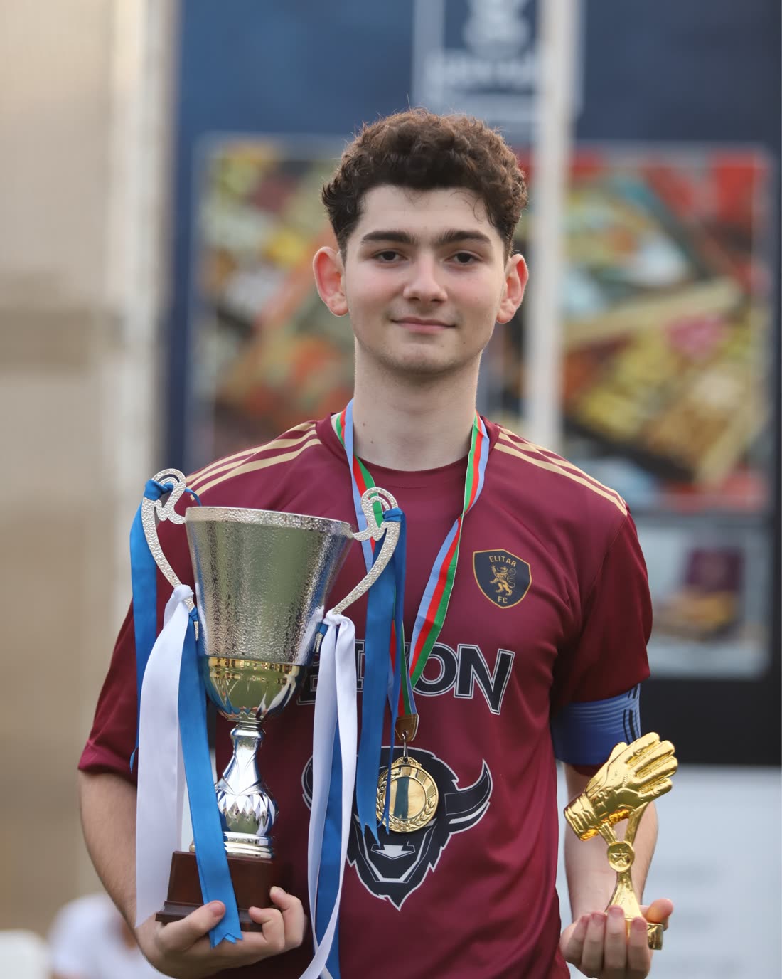 Teyyub in a maroon goalkeeper shirt holding a silver cup and a golden glove trophy